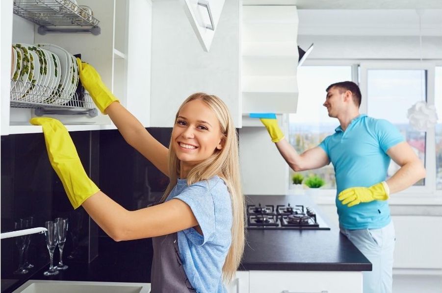 Two cleaners in a kitchen. A pretty blonde lady is stacking dishes and smiling. A man is cleaning in the background.