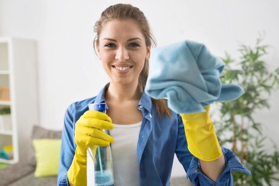 Lady cleaner holding cleaning spray and microfibre cloth. She is wearing a blue shirt and yellow rubber gloves. She is smiling.