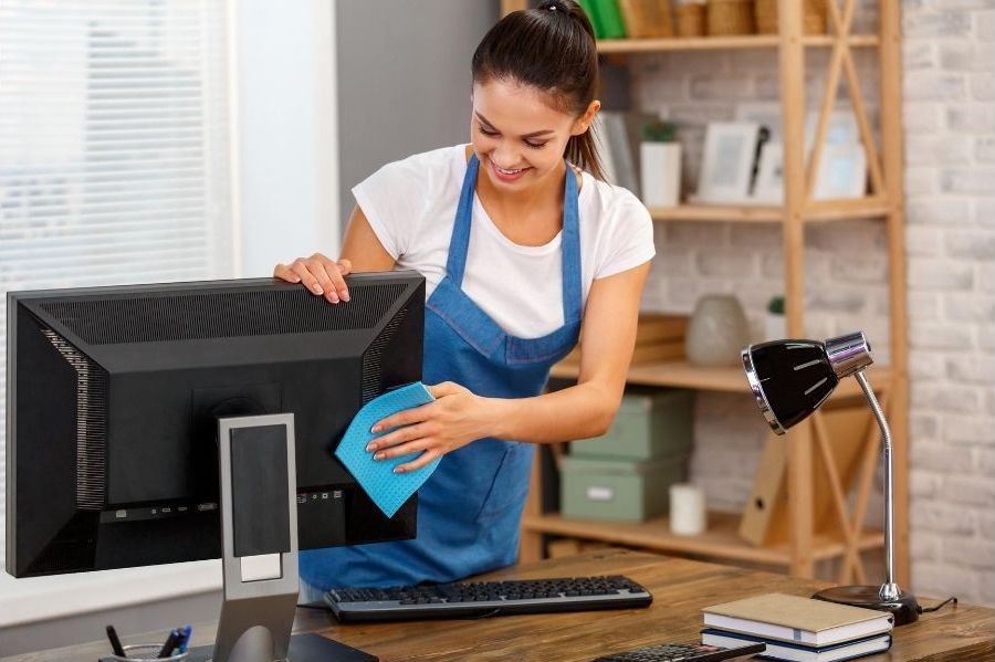 A lady cleaner is wiping a computer monitor with a blue cloth and smiling.